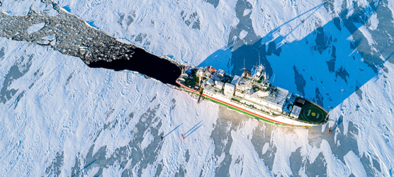 Ilmasta otettu kuva, alla merentutkimus alus halkoo jäätä, ihmisiä seisoo jäällä.